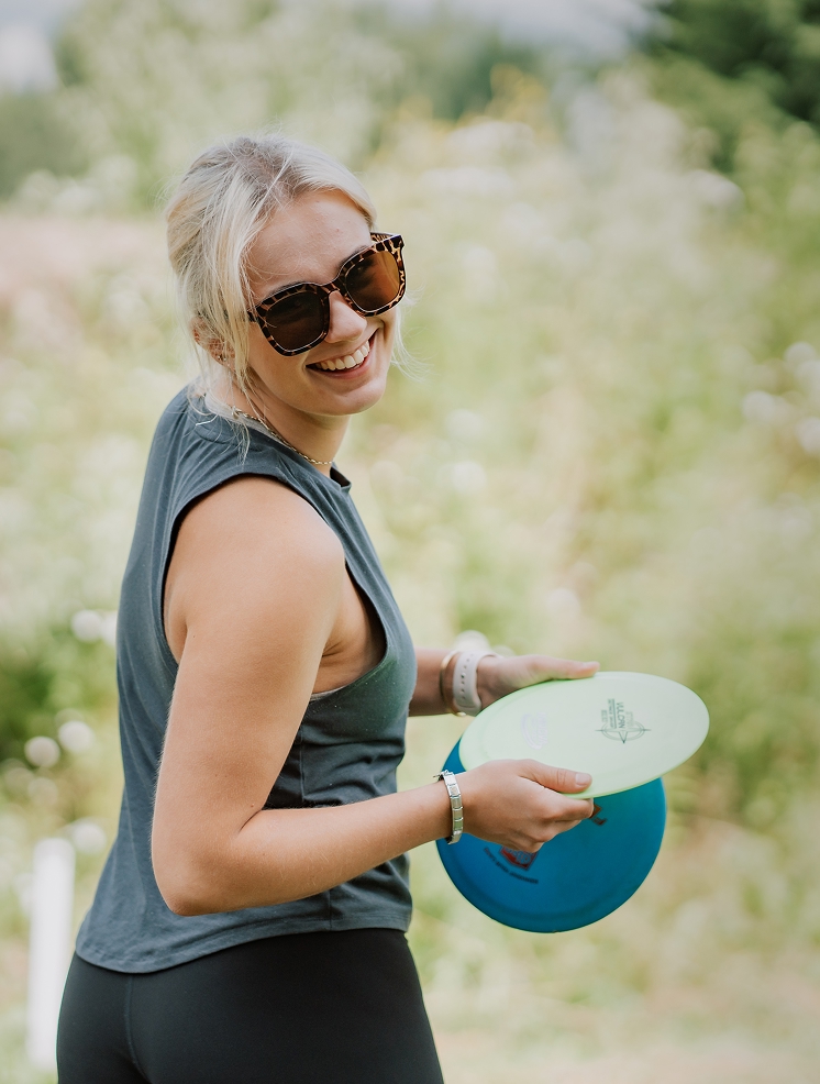 Smiling woman with sunglasses holding disc golf frisbees outdoors, enjoying recreational activity in a natural park setting.